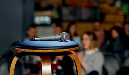 A microphone lies on a spotlit stool as a crowd gathers for a stand-up show at a local comedy club.